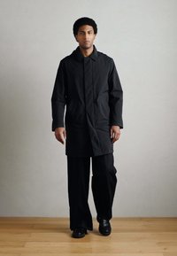 Fekete vízálló kabát egyenes szabással, galléros nyakkivágással, hosszú ujjakkal és enyhén texturált felülettel. Fekete nadrág fölött viselve.