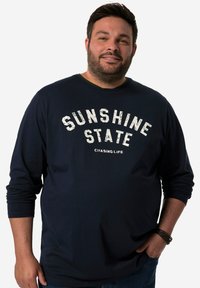 Námořnická modrá košile s dlouhým rukávem s nápisem "SUNSHINE STATE" vytištěným bílou barvou, s kulatým výstřihem a žebrovanými manžetami. V páru s džíny.