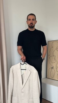 Ein hellbeiger Leinenblazer mit zwei Fronttaschen und einem Revers wird von einer Person in einem schwarzen Hemd an einem Kleiderbügel gehalten. Einfacher Hintergrund.