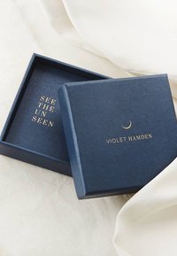 Marineblauwe vierkante geschenkdoos met gouden reliëftekst "ZIE HET ONGEZIENE" en "VIOLET HAMDEN"; matte afwerking, licht gestructureerd oppervlak, geopend deksel.