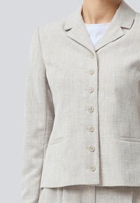 Světlý béžový blazer vyrobený z texturované látky, s klopovým límcem, šesti knoflíky na zapínání a dvěma předními kapsami.