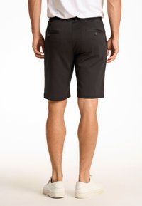 Schwarze Shorts aus einem glatten Stoff, mit zwei Gesäßtaschen und Knöpfen sowie einem geraden Schnitt, getragen mit weißen Sneakern.