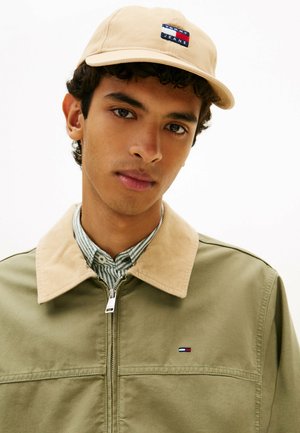 Joven con gorra beige de Tommy Jeans, chaqueta verde oliva con cuello beige y camisa de rayas abotonada, mirando ligeramente hacia un lado.