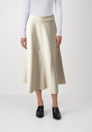 Žena nosiaca krémovú midi sukňu s panelovými švami, biely dlhý rukáv a čierne mules na podpätku, stojaca na neutrálnom pozadí.