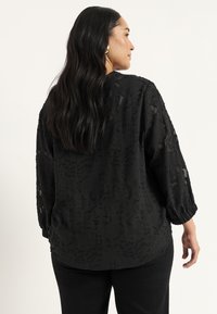 Sort, tekstureret bluse med en løs pasform, lange ærmer og indsnørede manchetter. Blødt stof med et subtilt mønster og rundet kant.