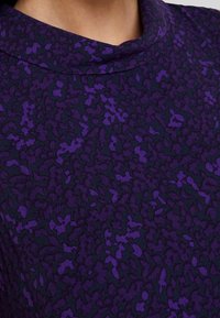 Fioletowa tkanina w wzory z mieszanką ciemnych i jasnych abstrakcyjnych kształtów. Widoczna miękka tekstura oraz wysoka linia dekoltu.