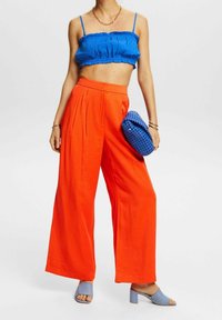Modrý zkrácený top s volánkovým detailem, spárovaný s jasně oranžovými kalhotami širokého střihu. Doplňky zahrnují texturovanou modrou kabelku a světlé modré lodičky.
