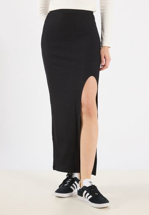 Femme portant une jupe noire taille haute avec une fente à la cuisse et des baskets Adidas noires et blanches sur fond blanc.