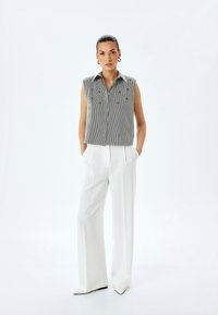 Camicia grigia senza maniche con bottoni, a righe verticali e ricami, abbinata a pantaloni bianchi a vita alta e scarpe bianche a punta.