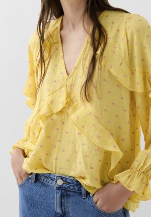 Femme portant un chemisier jaune à pois roses et volants, associé à un jean bleu, mains dans les poches, sur un fond uni.