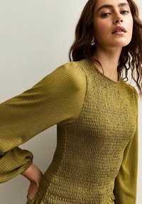 Top verde oliva con smock in rilievo, dotato di maniche lunghe e puffate e scollatura rotonda. Mette in evidenza un'intricata lavorazione e dettagli del tessuto.