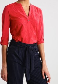 Blouse rouge texturé avec un décolleté en V et des manches trois-quarts, associé à un pantalon taille haute à rayures bleu marine et une ceinture assortie.