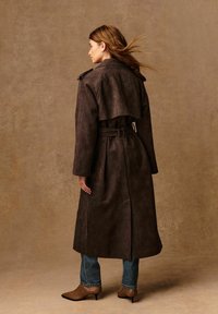 Cappotto trench in suede marrone con vita regolabile, ampio colletto e dettagli con patta; abbinato a jeans blu e stivali con tacco di colore neutro.