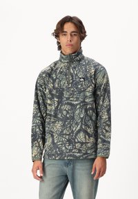 Fleece mikina s vysokým límcem, zdobená zeleným, černým a béžovým maskáčovým vzorem. Čtvrtzipový předek a logo nášivka na hrudi.