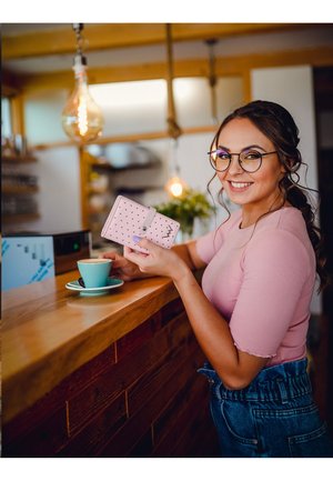 Pink wallet with white polka dots and a clasp, held near a turquoise coffee cup on a wooden counter. Bright setting with pendant lighting.