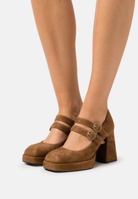 Brune ruskindssko med plateauhæl og runde spidser, ankelstropper med cirkulært metalbeslag og kraftige blokhæle. Glat tekstur.