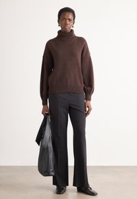 Maglione marrone oversize con collo a falda e orlo a coste, abbinato a pantaloni con righe verticali neri. Il modello tiene in mano una borsa di pelle nera.