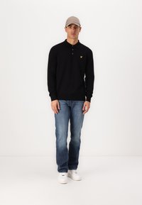 Svart långärmad polo skjorta med krage och gult logotyp på bröstet, i kombination med blå jeans och vita sneakers, bärande en ljusgrå keps.