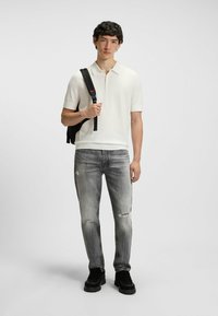 Hvid kortærmet polo, grå slidte jeans og sorte sneakers. Modellen bærer en sort rygsæk med minimalistisk design og glat tekstur.