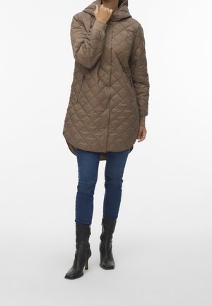 Kvinde iklædt en lang brun quiltet frakke med hætte, blå skinny jeans og sorte ankelstøvler med hæl, stående mod en hvid baggrund.