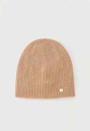 Bonnet beige en tricot côtelé avec un petit emblème rond doré près du bord inférieur sur un fond clair.