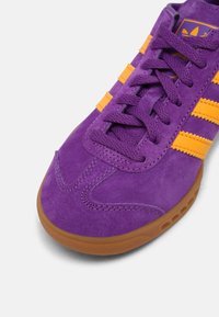 Purple suede sneaker with yellow stripes, featuring textured surface, round toe, and a gum rubber sole. Laces match the upper color.