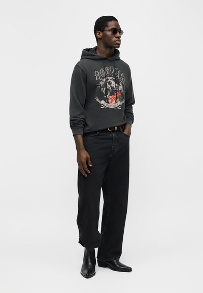 Άνδρας φορώντας μαύρο φούτερ με τύπωμα, μαύρο παντελόνι φαρδύ, μαύρες δερμάτινες μπότες, γυαλιά ηλίου, στέκεται με το ένα χέρι στην τσέπη μπροστά σε απλό φόντο.