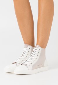 Bílé průsvitné vysoké tenisky s bílými šňůrkami obuté na nahých nohách na jednobarevném bílém pozadí.