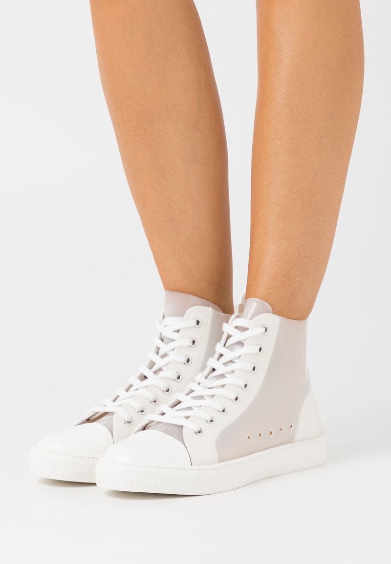 Bílé průsvitné vysoké tenisky s bílými šňůrkami obuté na nahých nohách na jednobarevném bílém pozadí.