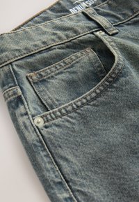 Gros plan sur une poche de jean bleu en denim avec une boucle de ceinture visible, des détails de couture et des rivets métalliques sur un tissu texturé.