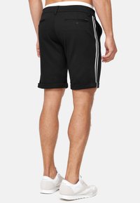 Schwarze, maßgeschneiderte Shorts aus glattem Stoff, mit umgeschlagenen Säumen und weißen Seitenstreifen. Enthält Gesäßtaschen und einen Knopfdetails.