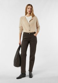 Vrouw staand, draagt een beige vest met knopen over een witte top, donkere broek, zwarte enkelboots, en houdt een grote zwarte geweven tas vast.