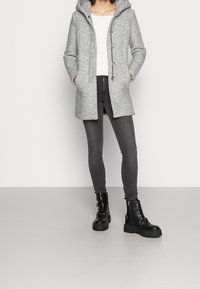 Šedý, kapucí zdobený plášť se strukturovaným povrchem, v kombinaci s přiléhavým bílým svetr, tmavě šedými džíny a černými robustními botami s přezkami.