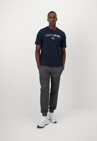 Camisa azul-marinho de manga curta com logotipo branco, combinada com calças jogger cinzas e ténis brancos. O conjunto apresenta um corte descontraído e um design casual.
