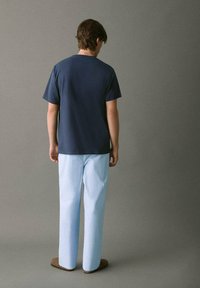 T-paita tummansinisellä lyhyellä hihaisella, yhdistettynä vaaleansinisiin raidallisiin housuihin ja ruskeisiin liukuvaan tyyliin sandaaleihin, nähtynä selkäpuolelta.