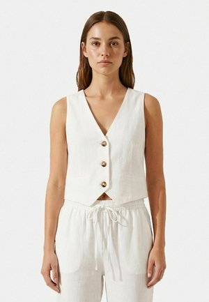 Kobieta ubrana w białą kamizelkę bez rękawów z trzema guzikami oraz białe spodnie z troczkami, stojąca na jednolitym jasnym tle.