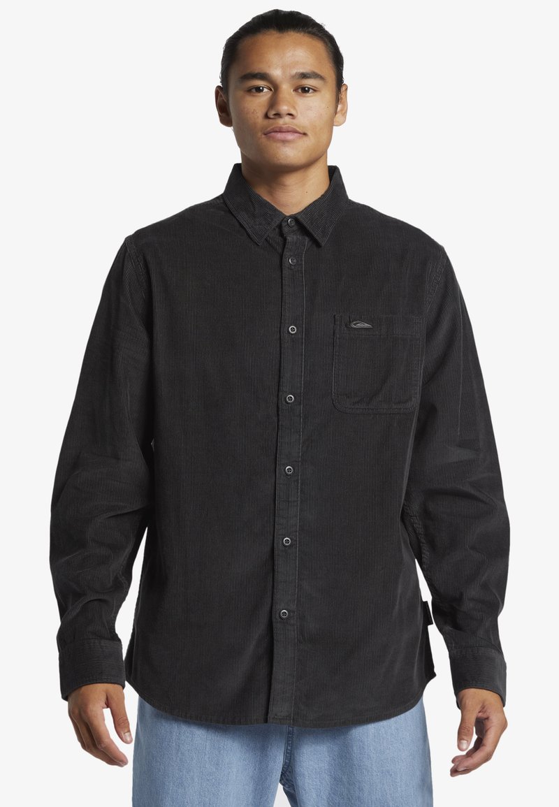 Camisa de pana negra con mangas largas, cierre de botones en la parte delantera, un bolsillo en el pecho y una textura sutil. Combinada con jeans de color azul claro.