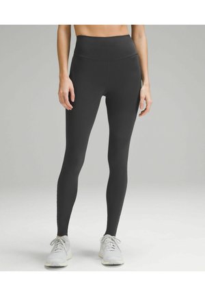 Frau trägt schwarze, hochtaillierte Leggings in voller Länge und weiße Sportschuhe, steht vor einem schlichten hellgrauen Hintergrund.