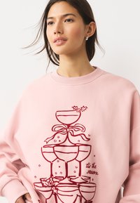 Svetloružová mikina s červeným obrysom navrstvených kokteilových pohárov s cesmínou a mašľou, spolu s textom „tis the season“.