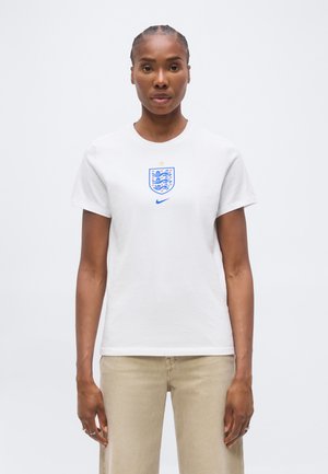 Vrouw met gevlochten haar die een wit t-shirt van het Engelse voetbalteam en beige broek draagt, staand tegen een effen witte achtergrond.