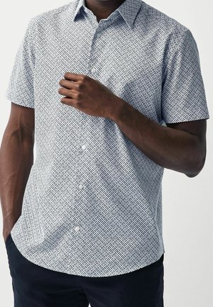 Homme portant une chemise à manches courtes boutonnée avec un motif géométrique en losanges noirs et blancs, une main ajustant le col, l'autre dans la poche.