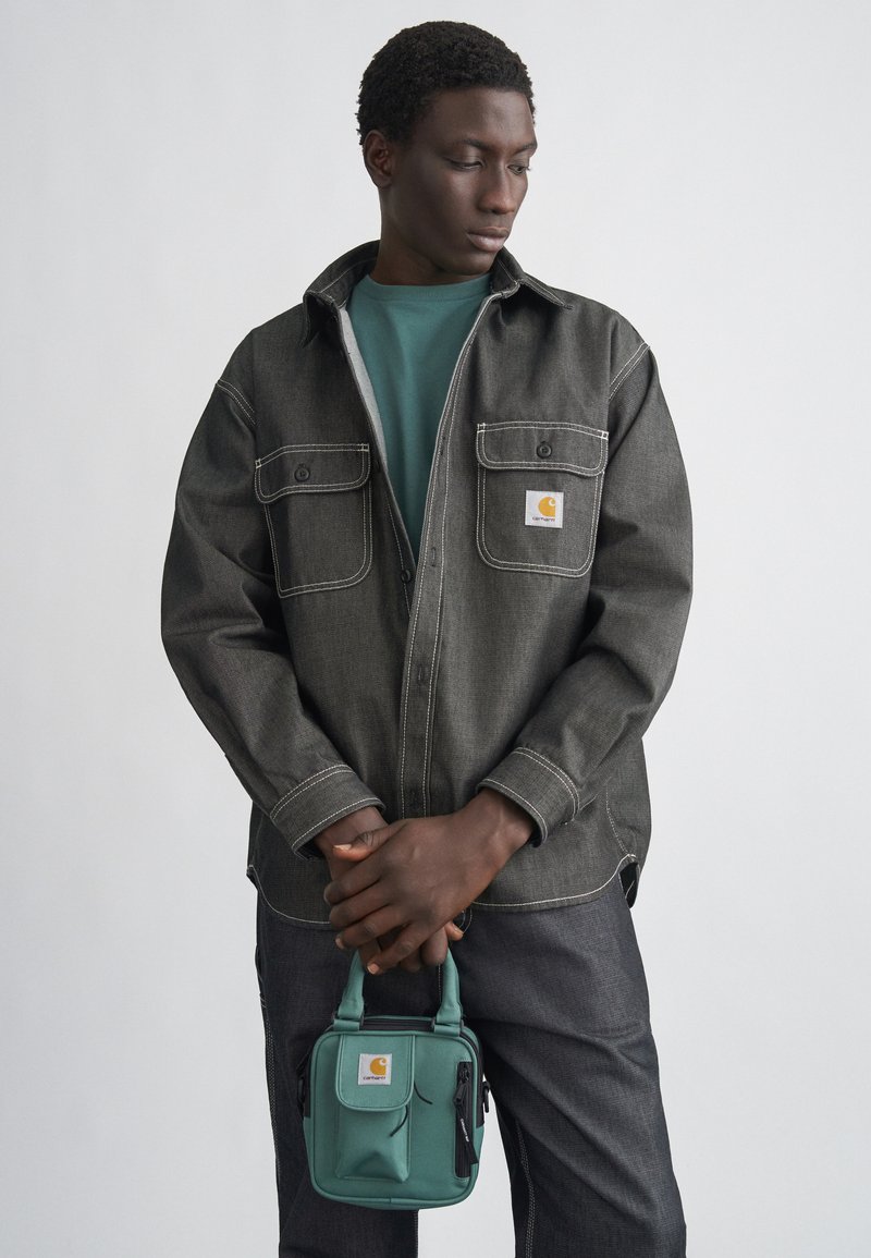 Czarna koszula jeansowa z szarym przeszyciem, dwoma kieszeniami na piersi i logo. Pod spodem morska koszulka, ciemne spodnie oraz morska torba na ramię z zamkami błyskawicznymi.