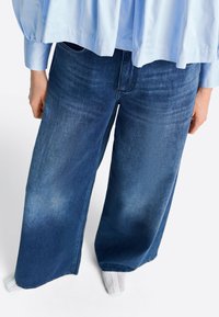 Közép kék farmerszínű bőszárú nadrág, kopott hatással. Világoskék, lazán illeszkedő inggel párosítva; látható fehér mintás zoknik.