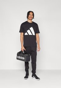 Juodas marškinėliai su dideliu baltu Adidas logotipu, juodos sportinės kelnės, juodi sportbačiai ir juodas sportinis krepšys su baltu Adidas ženklu.