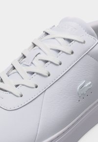 Κοντινό πλάνο λευκού δερμάτινου αθλητικού παπουτσιού με λευκά κορδόνια και μικρό λογότυπο κροκόδειλου στο πλάι κοντά στην σόλα.