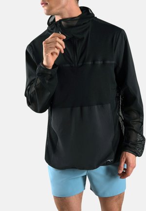 Man in zwart gaasjack met rits, kraag verstelend, draagt lichtblauwe korte broek, staand tegen een witte achtergrond.