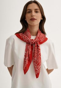 Czerwony szalik w wzór paisley, wykonany z lekkiego materiału, związany wokół szyi. Nosi się go na luźnej białej bluzce. Gładka faktura.