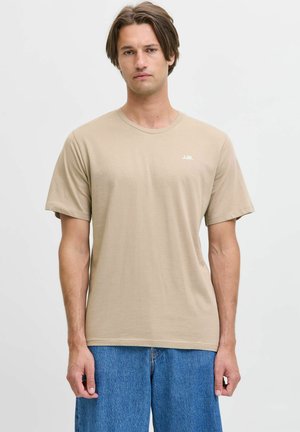 Joven con cabello castaño que lleva una camiseta beige de manga corta y vaqueros azules, de pie frente a un fondo liso y claro.