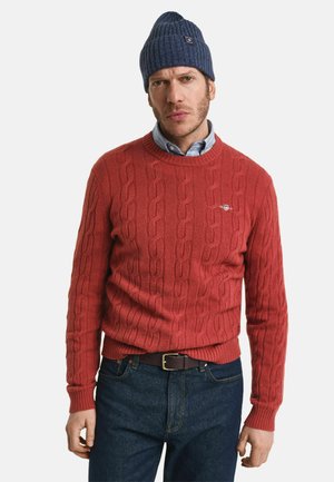 Roter Kabelstrickpullover mit rundem Halsausschnitt, kombiniert mit einer marineblauen Rippmütze. Trägt man über einem hellblauen Hemd mit Kragen und dunklen Jeans.