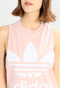 Ung kvinna som bär en ärmlös rosa topp med en stor vit Adidas-logotyp, beskuren vid axlarna och munnen, mot en enkel bakgrund.